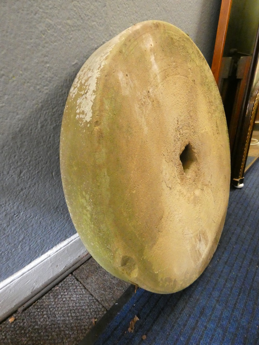 A circular hand-carved stone millstone featuring a square central aperture, concentric grooved circles, and a weathered moss-covered surface. View III