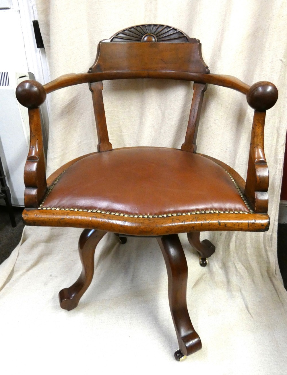 An antique revolving wood desk chair featuring a carved sunburst crest rail, curved armrests, a brown leather seat with brass stud trimming, and splayed legs on castors.