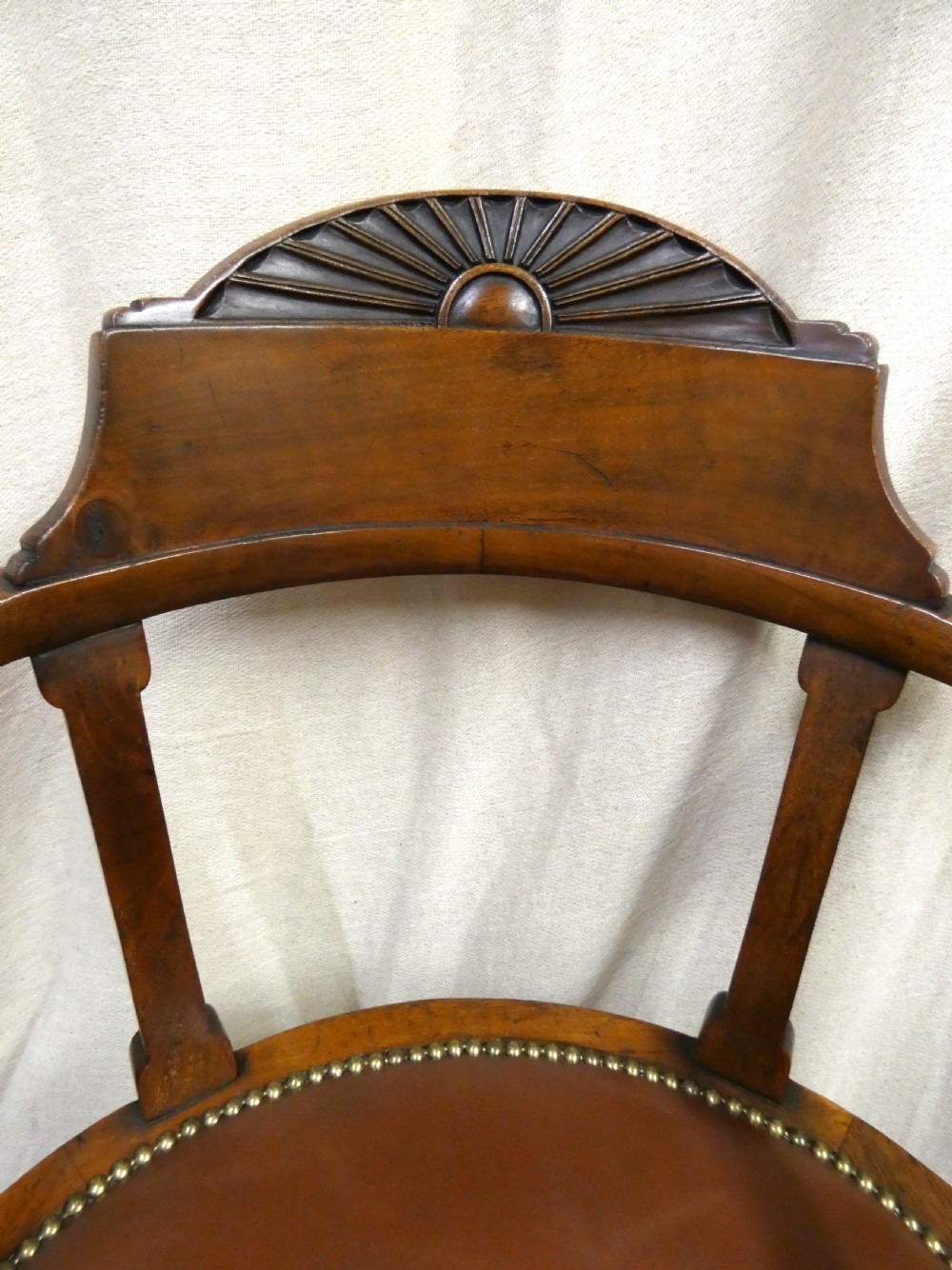 An antique revolving wood desk chair featuring a carved sunburst crest rail, curved armrests, a brown leather seat with brass stud trimming, and splayed legs on castors. View II