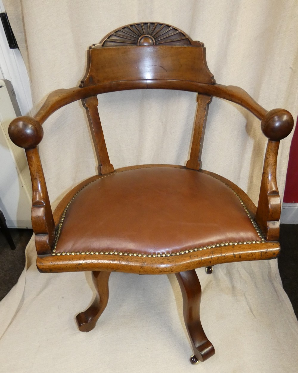 An antique revolving wood desk chair featuring a carved sunburst crest rail, curved armrests, a brown leather seat with brass stud trimming, and splayed legs on castors. View III