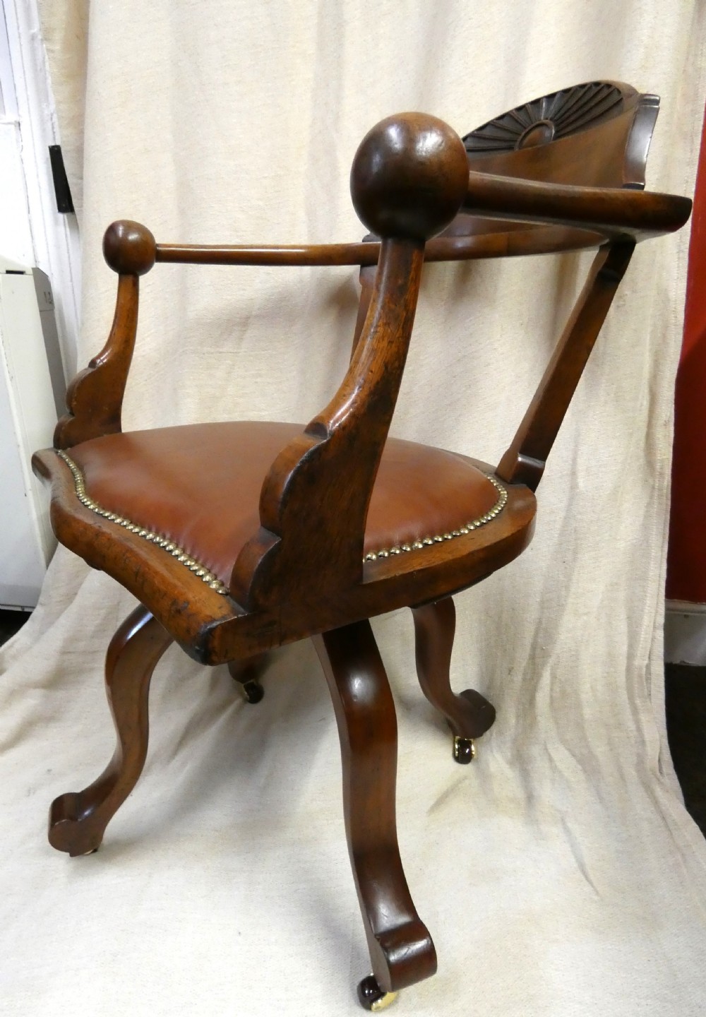 An antique revolving wood desk chair featuring a carved sunburst crest rail, curved armrests, a brown leather seat with brass stud trimming, and splayed legs on castors. View VI