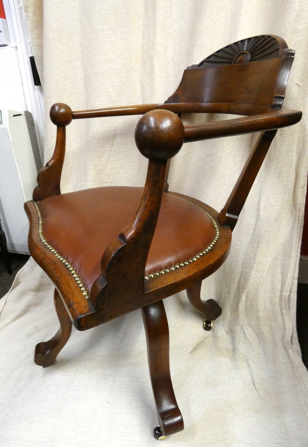 An antique revolving wood desk chair featuring a carved sunburst crest rail, curved armrests, a brown leather seat with brass stud trimming, and splayed legs on castors. View VII