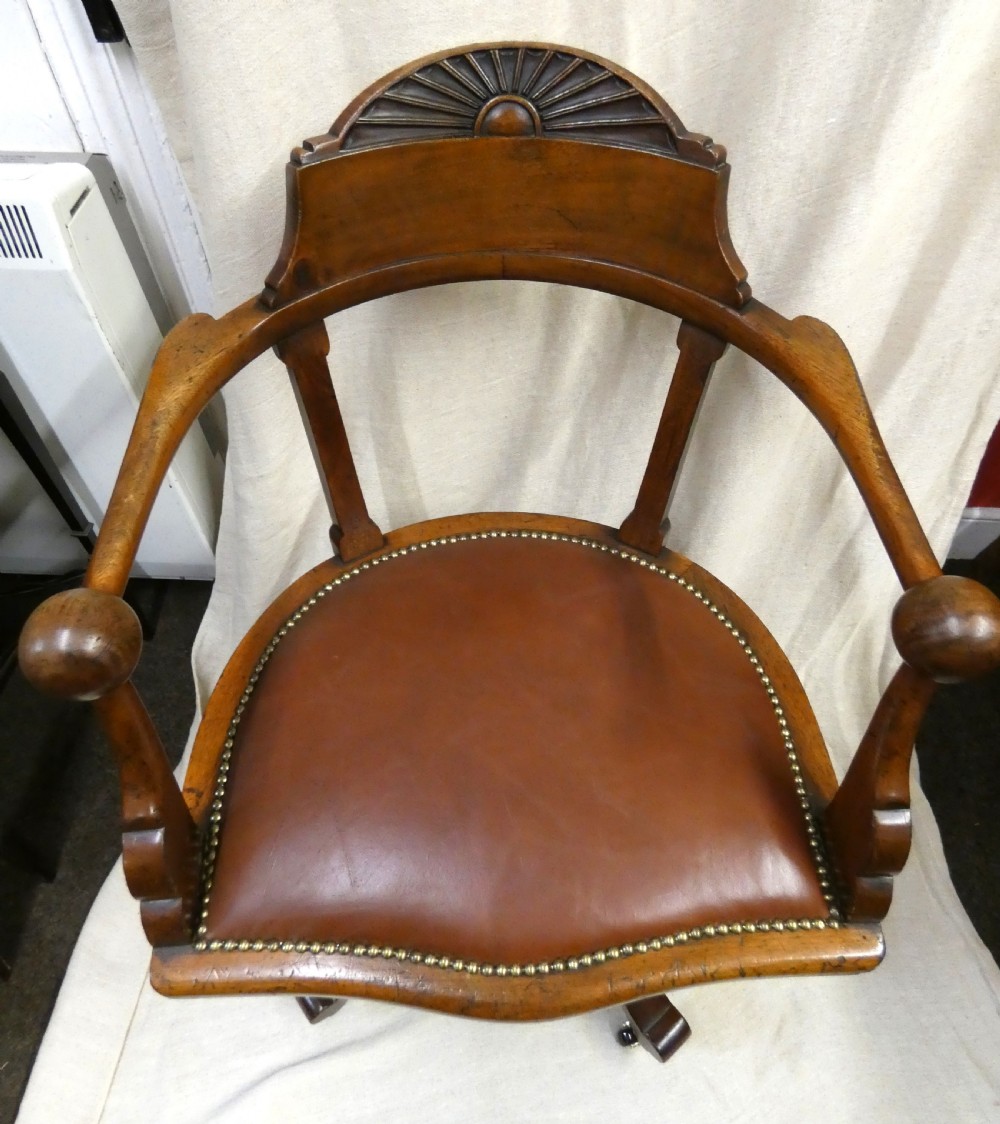 An antique revolving wood desk chair featuring a carved sunburst crest rail, curved armrests, a brown leather seat with brass stud trimming, and splayed legs on castors. View VIII