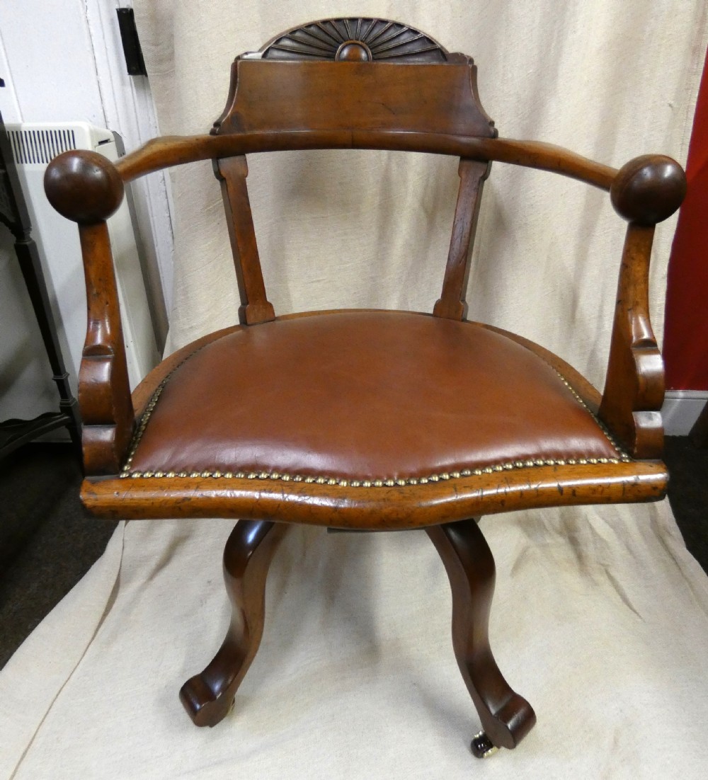 An antique revolving wood desk chair featuring a carved sunburst crest rail, curved armrests, a brown leather seat with brass stud trimming, and splayed legs on castors. View IX