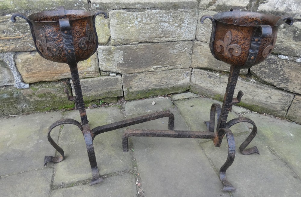 A pair of wrought iron andirons featuring scrolled legs, upright stems, and hammered copper basin tops decorated with fleur-de-lis motifs.