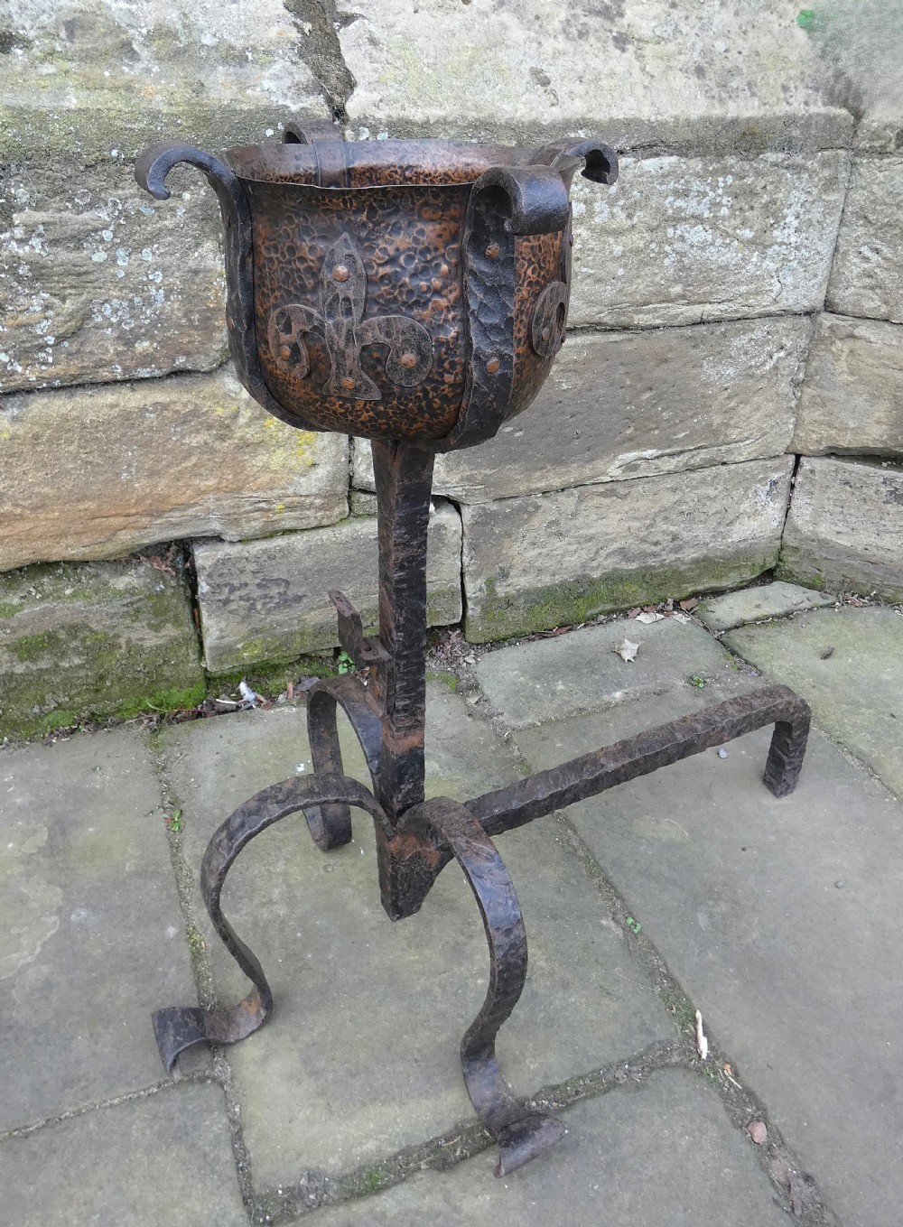A pair of wrought iron andirons featuring scrolled legs, upright stems, and hammered copper basin tops decorated with fleur-de-lis motifs. View II
