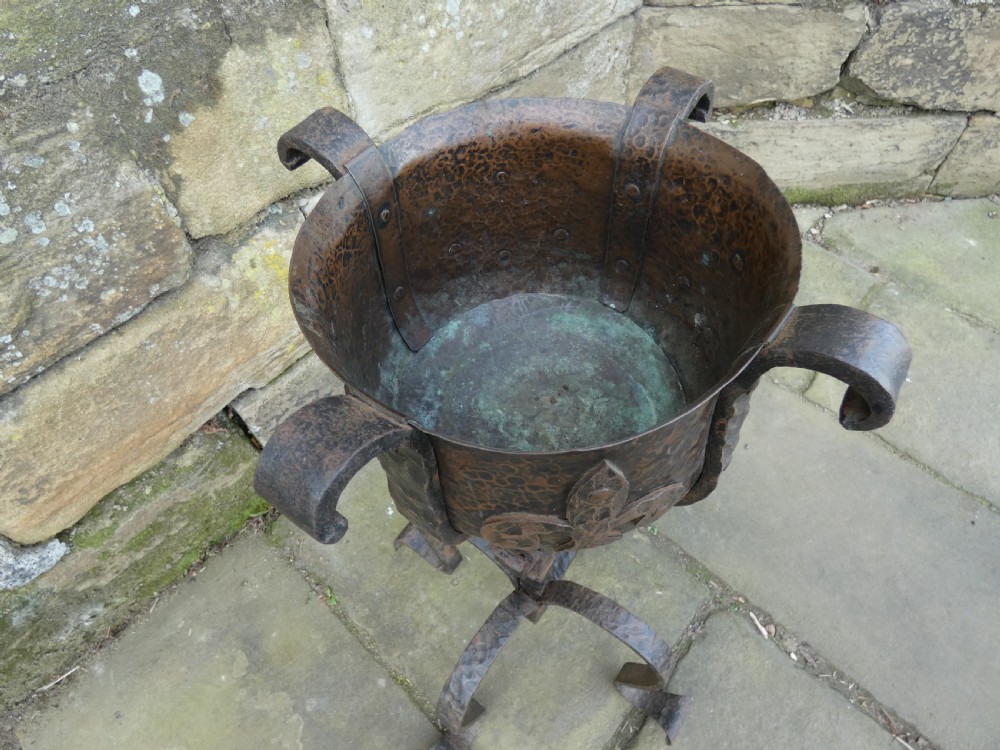 A pair of wrought iron andirons featuring scrolled legs, upright stems, and hammered copper basin tops decorated with fleur-de-lis motifs. View X