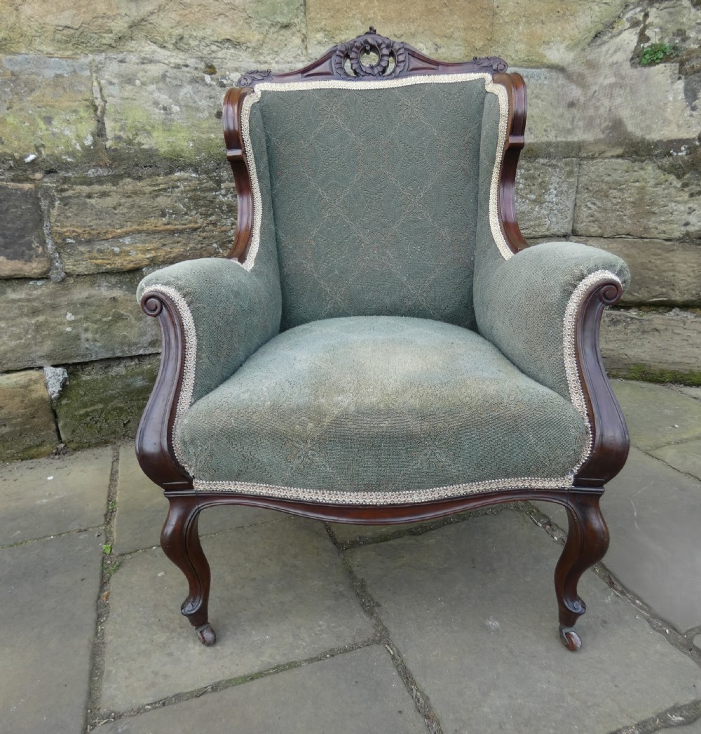 An antique Victorian mahogany armchair featuring sage green patterned fabric upholstery, decorative braid trim, a carved crest rail, and cabriole front legs on brass castors.
