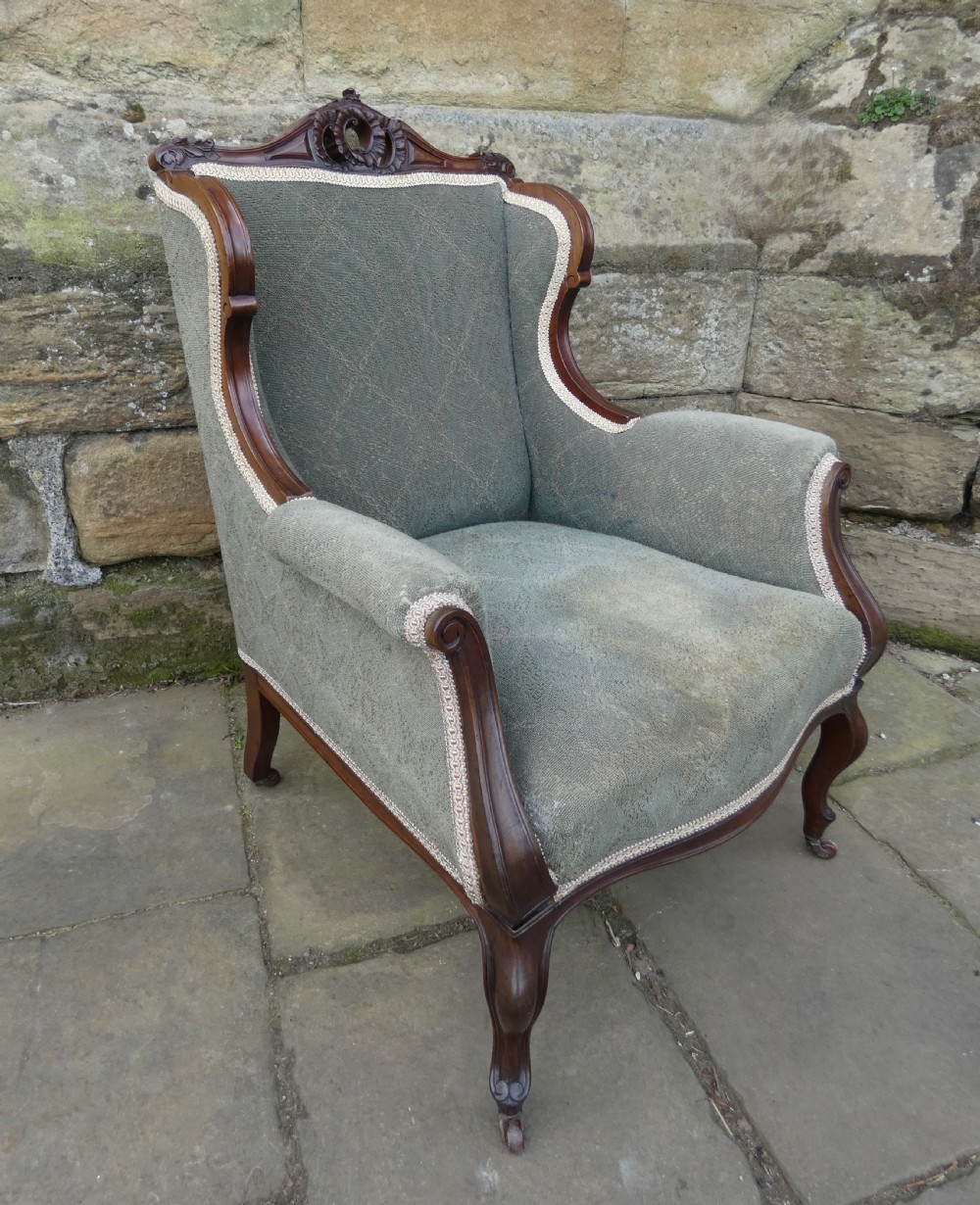 An antique Victorian mahogany armchair featuring sage green patterned fabric upholstery, decorative braid trim, a carved crest rail, and cabriole front legs on brass castors. View II