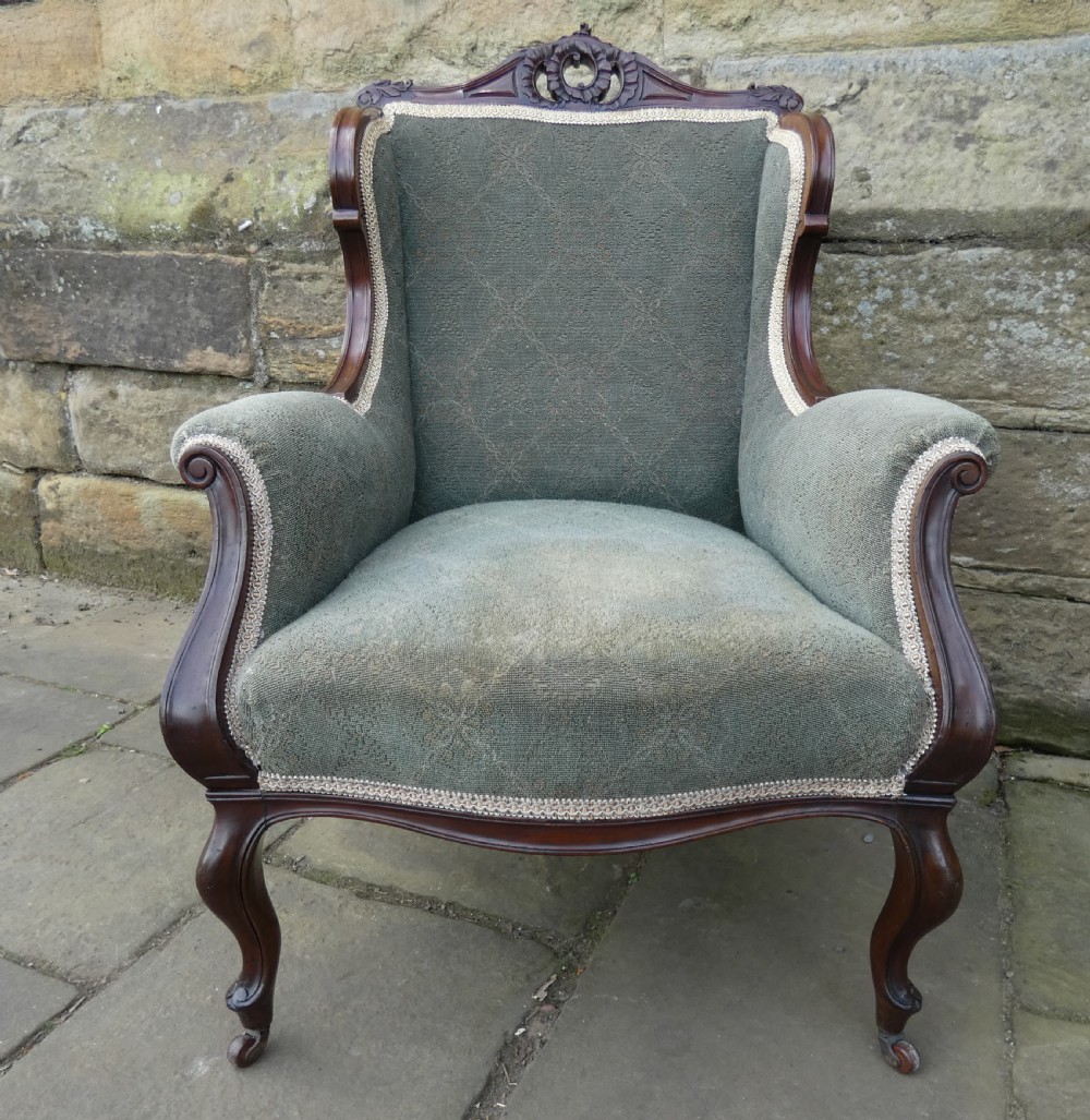 An antique Victorian mahogany armchair featuring sage green patterned fabric upholstery, decorative braid trim, a carved crest rail, and cabriole front legs on brass castors. View III