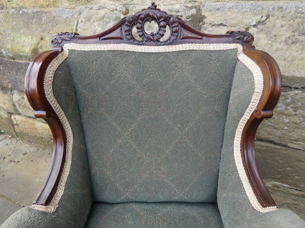 An antique Victorian mahogany armchair featuring sage green patterned fabric upholstery, decorative braid trim, a carved crest rail, and cabriole front legs on brass castors. View V