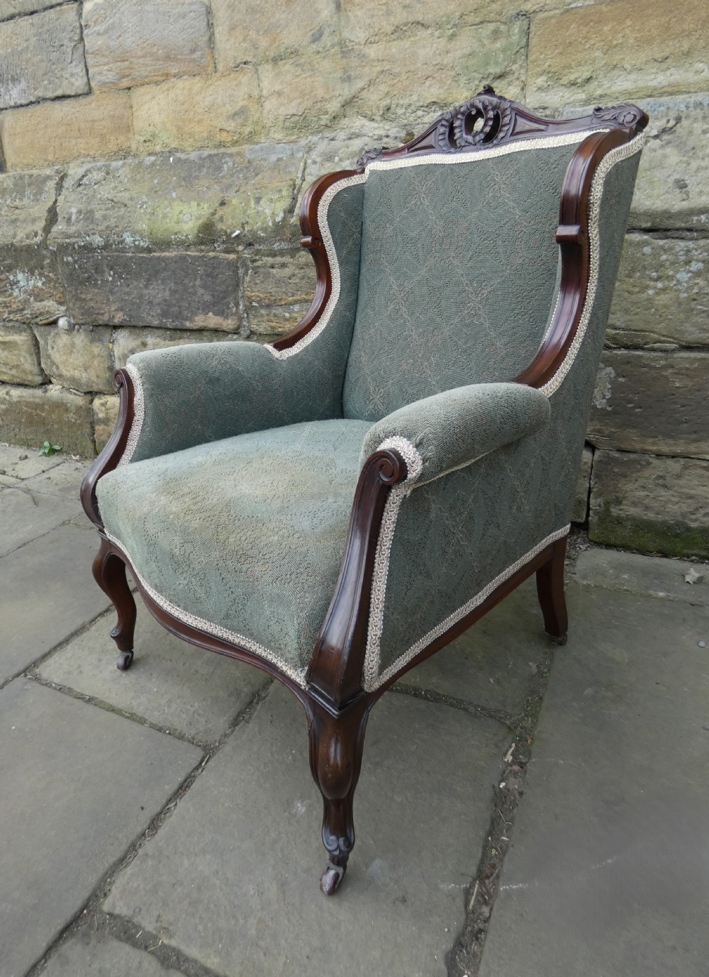 An antique Victorian mahogany armchair featuring sage green patterned fabric upholstery, decorative braid trim, a carved crest rail, and cabriole front legs on brass castors. View VI