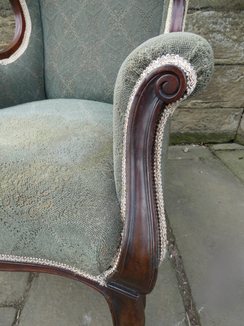 An antique Victorian mahogany armchair featuring sage green patterned fabric upholstery, decorative braid trim, a carved crest rail, and cabriole front legs on brass castors. View VII