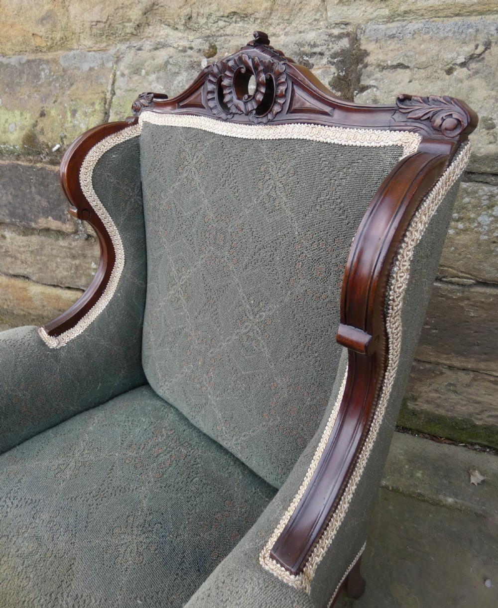 An antique Victorian mahogany armchair featuring sage green patterned fabric upholstery, decorative braid trim, a carved crest rail, and cabriole front legs on brass castors. View VIII
