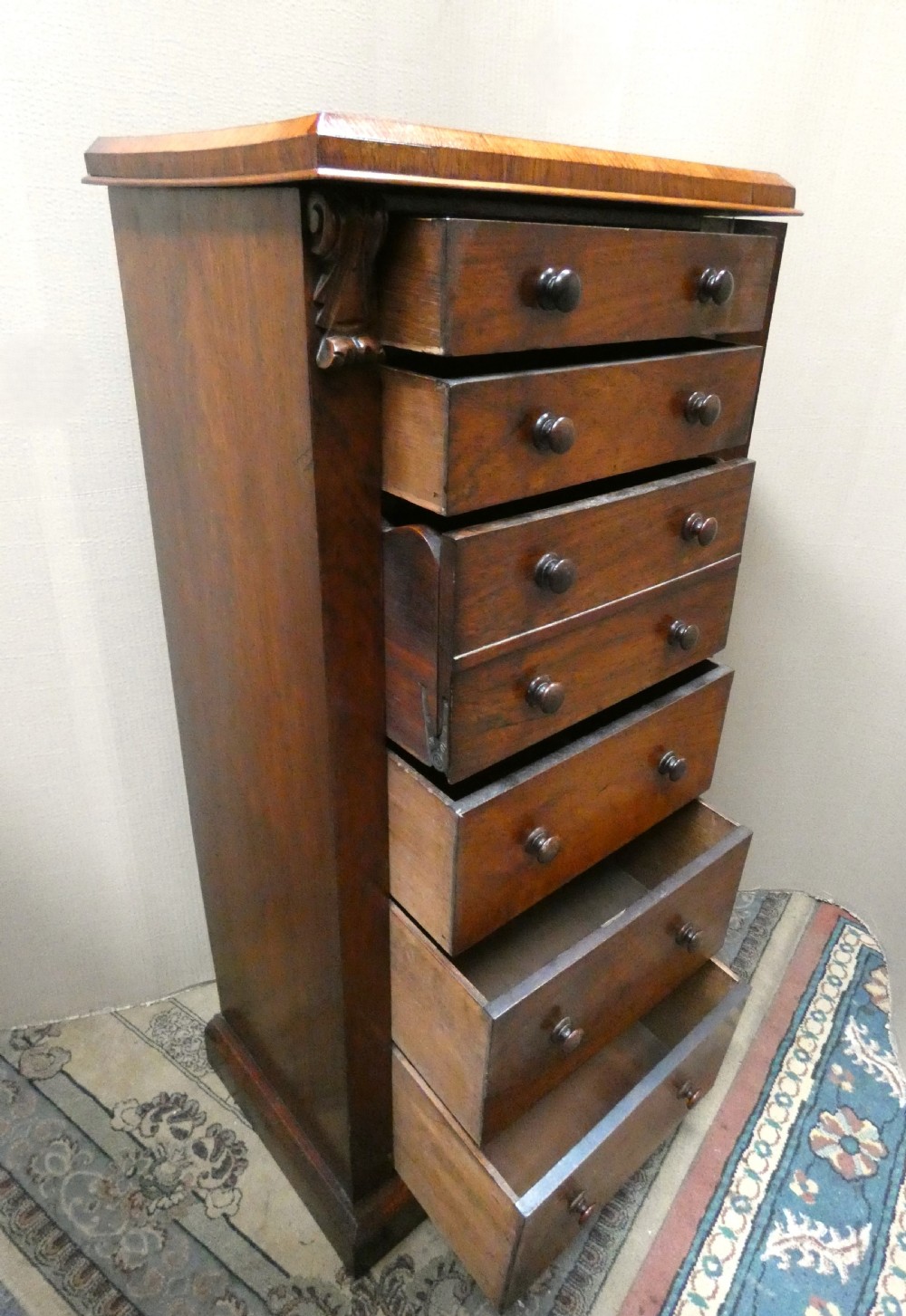 A tall Victorian rosewood Wellington chest featuring turned wooden knobs, carved upper corbels, and an open drop-front secretaire drawer revealing internal pigeonholes and small drawers. View VIII