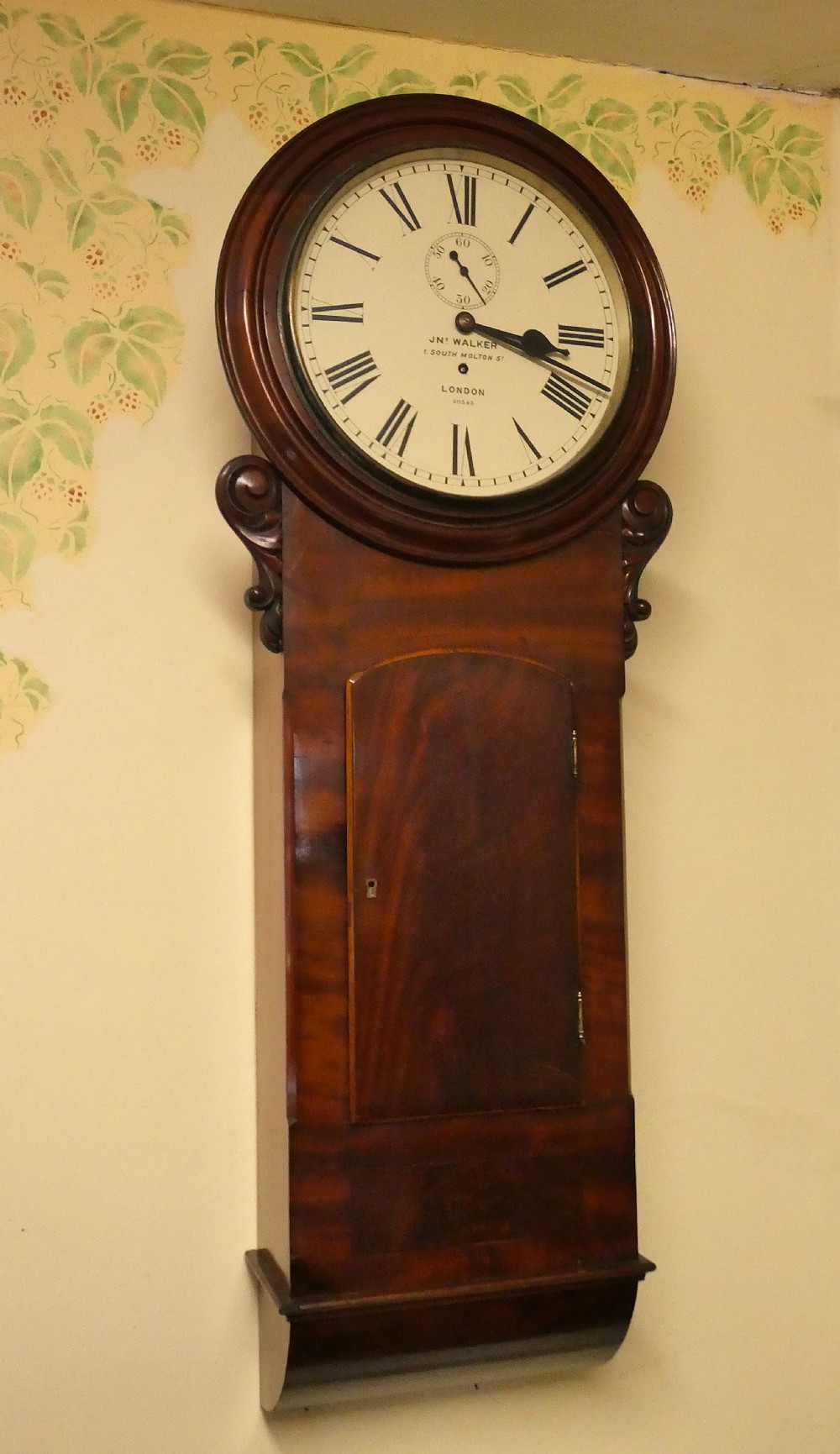 A Victorian flame mahogany regulator wall clock featuring a round white dial with Roman numerals above a drop trunk case with carved scrolls.