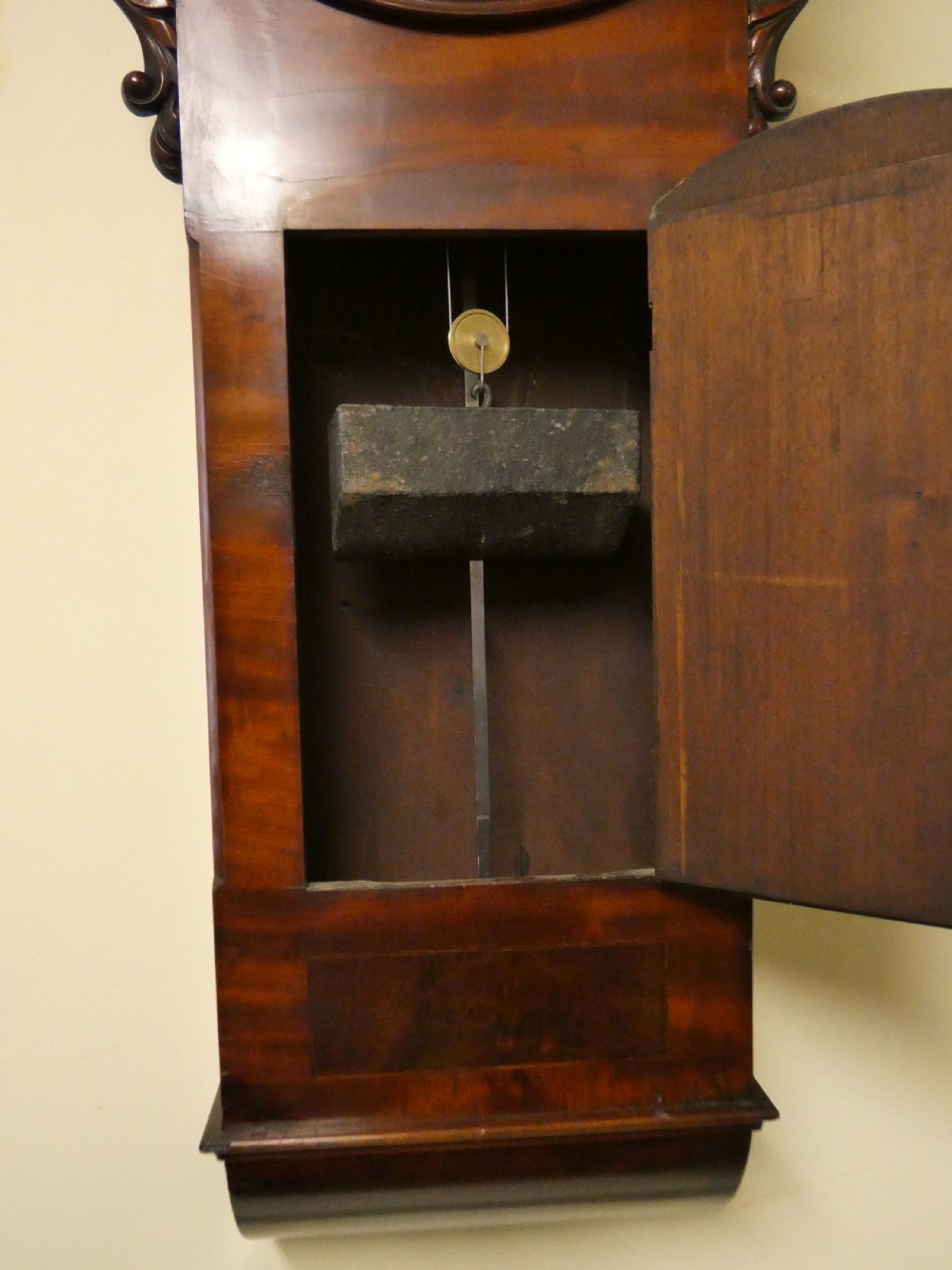 A Victorian flame mahogany regulator wall clock featuring a round white dial with Roman numerals above a drop trunk case with carved scrolls. View VI