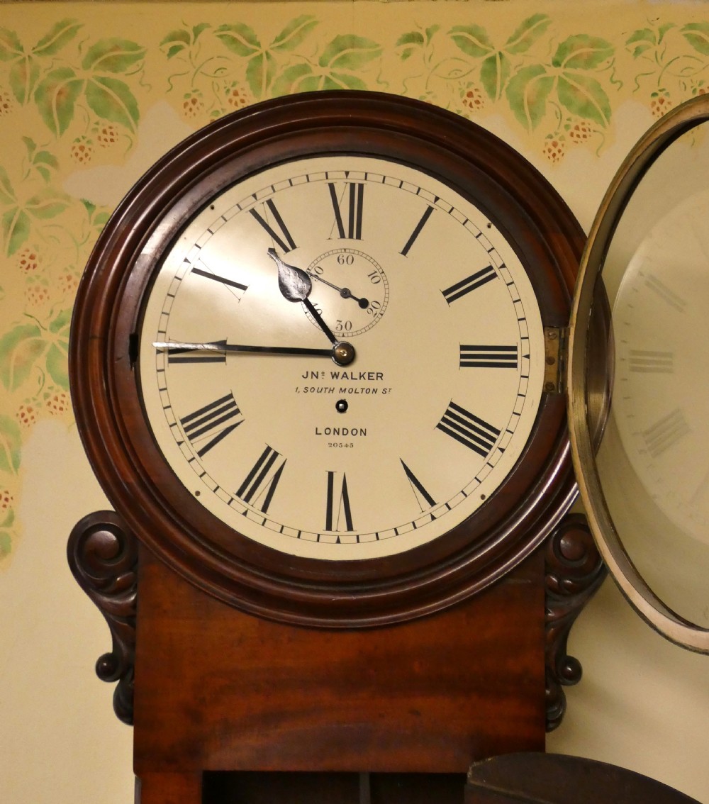 A Victorian flame mahogany regulator wall clock featuring a round white dial with Roman numerals above a drop trunk case with carved scrolls. View VII