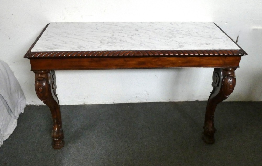 A William IV mahogany console table featuring a light marble top, gadrooned apron rim, and carved scroll front legs.