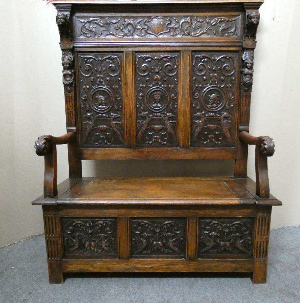 A dark carved oak high-back settle featuring three relief-carved back panels with portrait medallions, side caryatid figures, and a hinged storage seat above carved base panels.