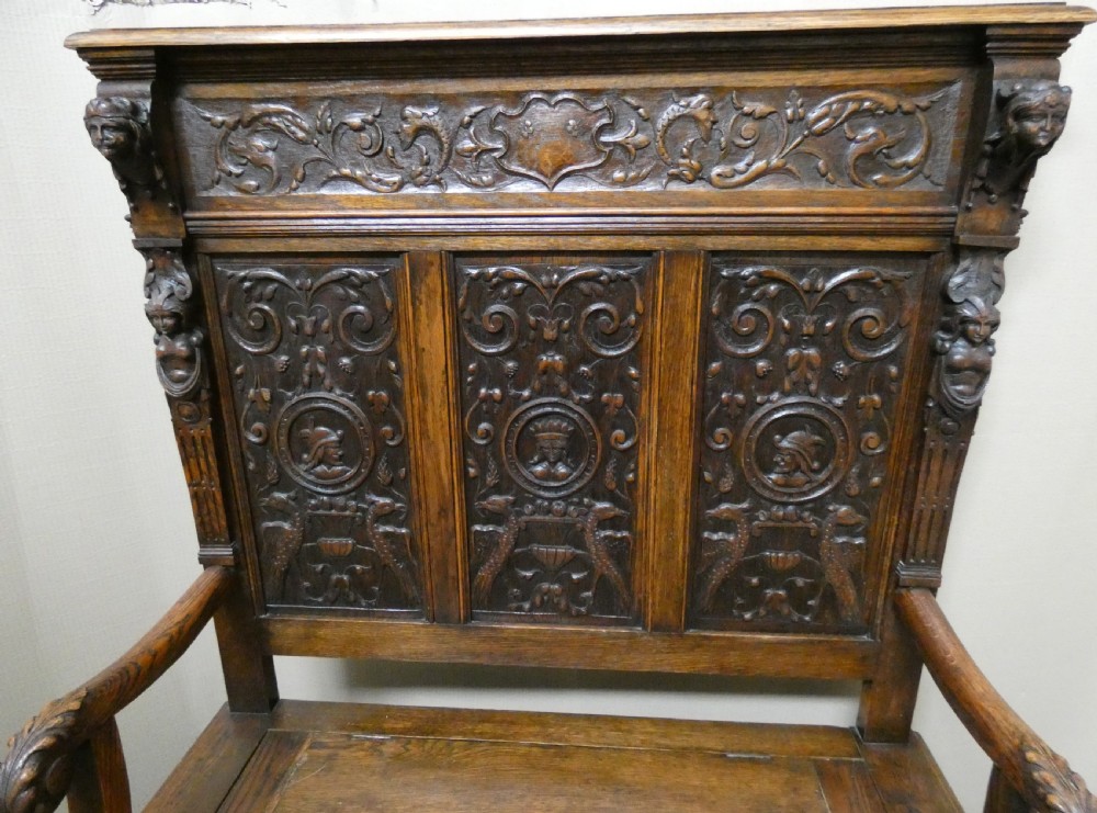 A dark carved oak high-back settle featuring three relief-carved back panels with portrait medallions, side caryatid figures, and a hinged storage seat above carved base panels. View II