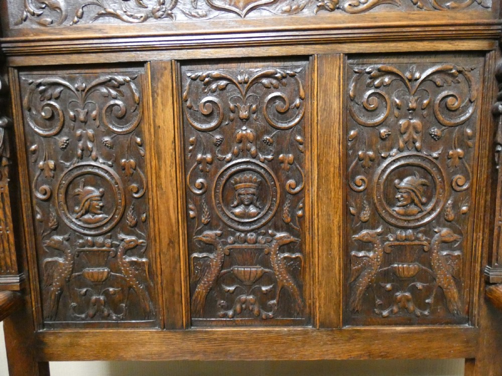 A dark carved oak high-back settle featuring three relief-carved back panels with portrait medallions, side caryatid figures, and a hinged storage seat above carved base panels. View III