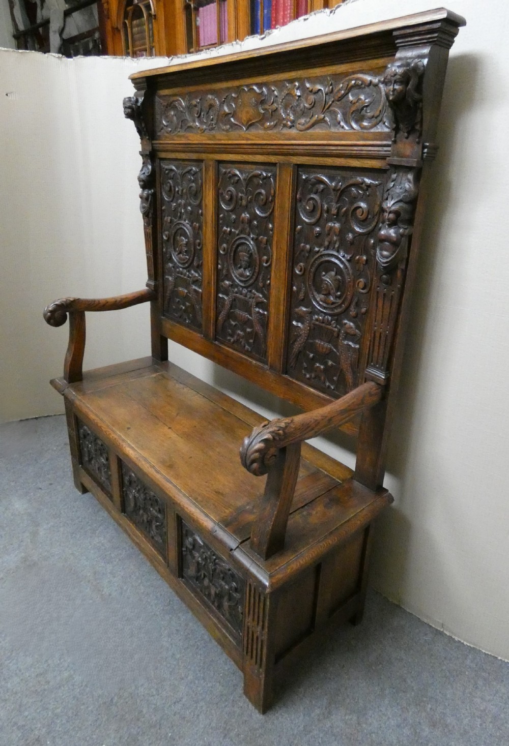 A dark carved oak high-back settle featuring three relief-carved back panels with portrait medallions, side caryatid figures, and a hinged storage seat above carved base panels. View X
