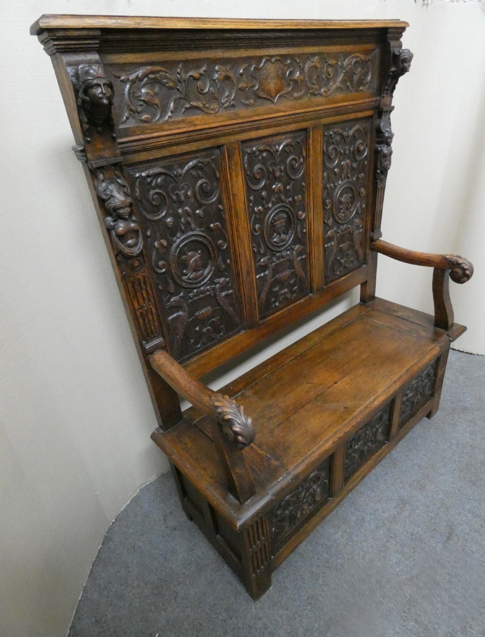 A dark carved oak high-back settle featuring three relief-carved back panels with portrait medallions, side caryatid figures, and a hinged storage seat above carved base panels. View XI