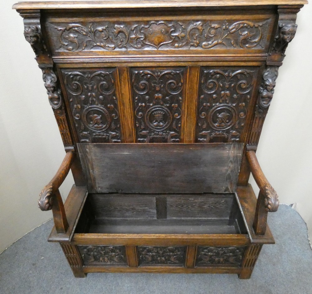 A dark carved oak high-back settle featuring three relief-carved back panels with portrait medallions, side caryatid figures, and a hinged storage seat above carved base panels. View XII
