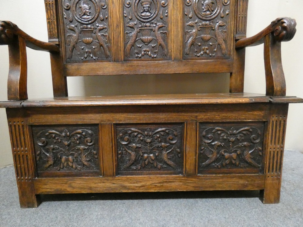 A dark carved oak high-back settle featuring three relief-carved back panels with portrait medallions, side caryatid figures, and a hinged storage seat above carved base panels. View XIII