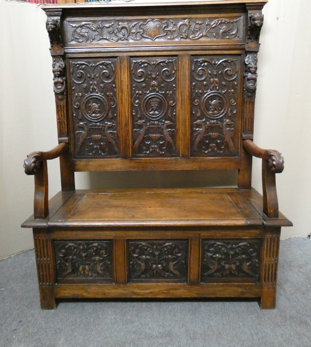 A dark carved oak high-back settle featuring three relief-carved back panels with portrait medallions, side caryatid figures, and a hinged storage seat above carved base panels. View XIV