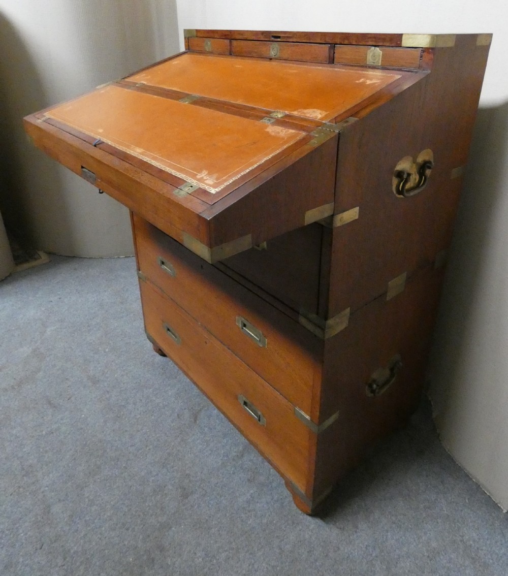 An antique teak campaign secretaire chest featuring brass corner mounts, flush drawer handles, carrying handles, and a drop-front slanted leather writing desk.