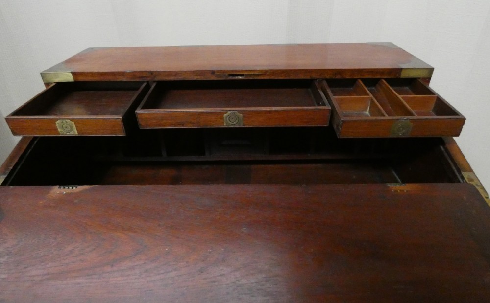 An antique teak campaign secretaire chest featuring brass corner mounts, flush drawer handles, carrying handles, and a drop-front slanted leather writing desk. View V
