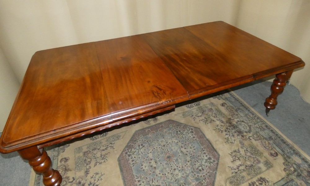A rectangular mahogany extending dining table with two removable leaves displayed upright, resting on heavy turned legs with brass castors. View III