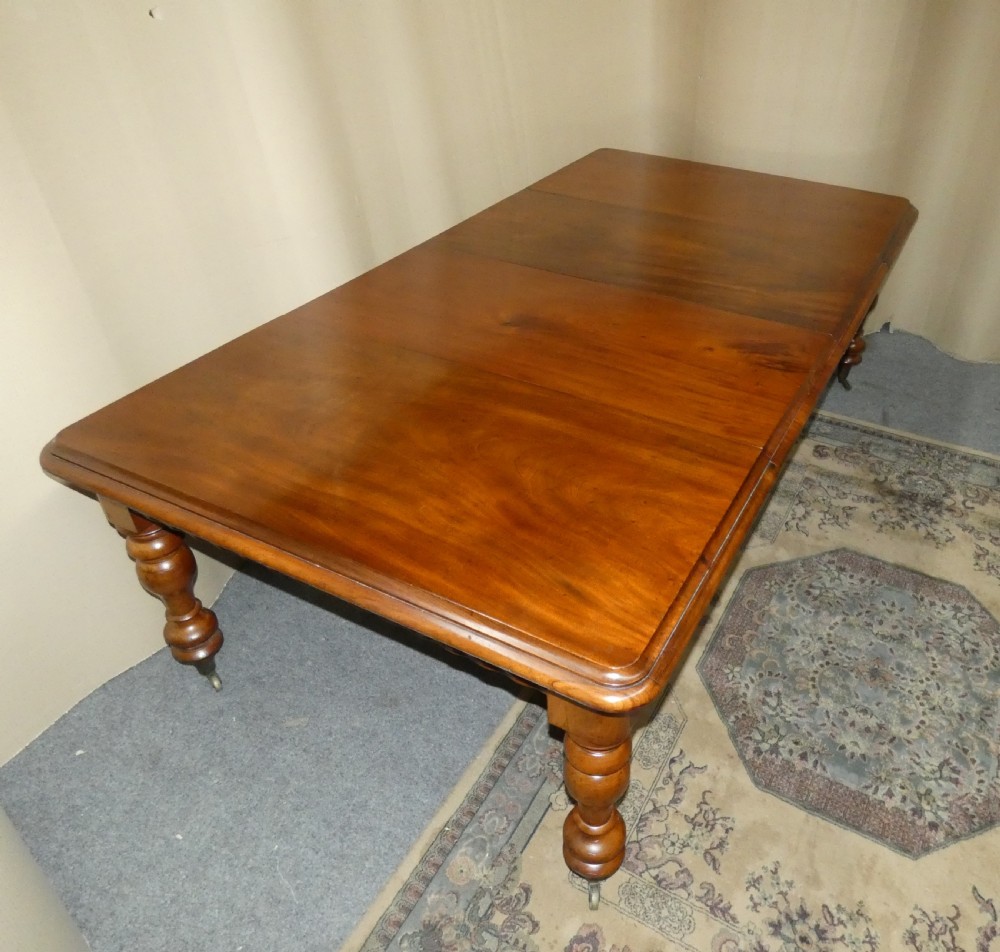 A rectangular mahogany extending dining table with two removable leaves displayed upright, resting on heavy turned legs with brass castors. View V