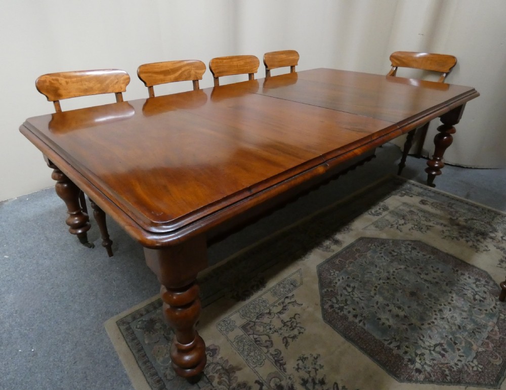A rectangular mahogany extending dining table with two removable leaves displayed upright, resting on heavy turned legs with brass castors. View VII