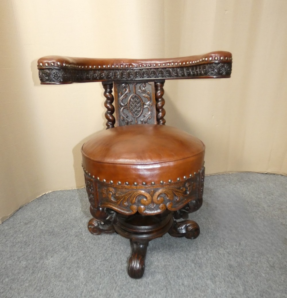 A carved dark oak cockfighting chair featuring tan leather upholstery with brass stud trimmings, a wide padded armrest, and barley twist support posts.