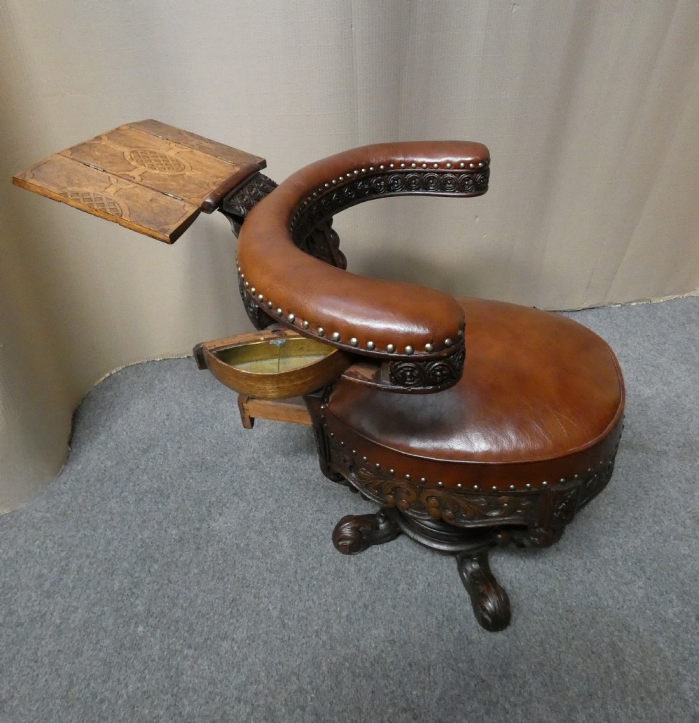 A carved dark oak cockfighting chair featuring tan leather upholstery with brass stud trimmings, a wide padded armrest, and barley twist support posts. View II