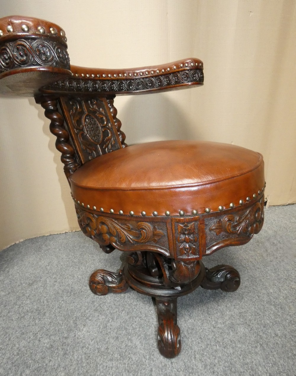 A carved dark oak cockfighting chair featuring tan leather upholstery with brass stud trimmings, a wide padded armrest, and barley twist support posts. View V