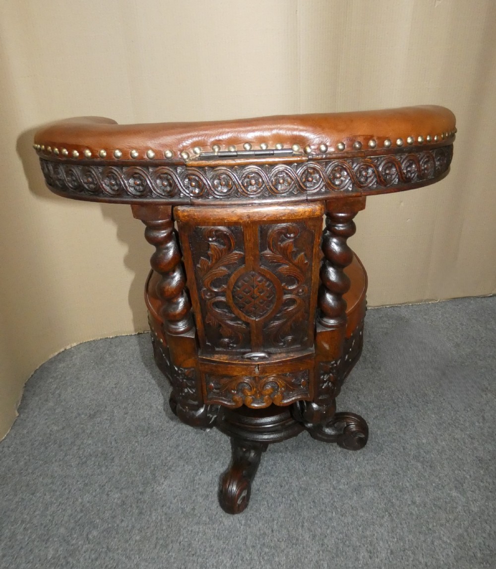A carved dark oak cockfighting chair featuring tan leather upholstery with brass stud trimmings, a wide padded armrest, and barley twist support posts. View IX