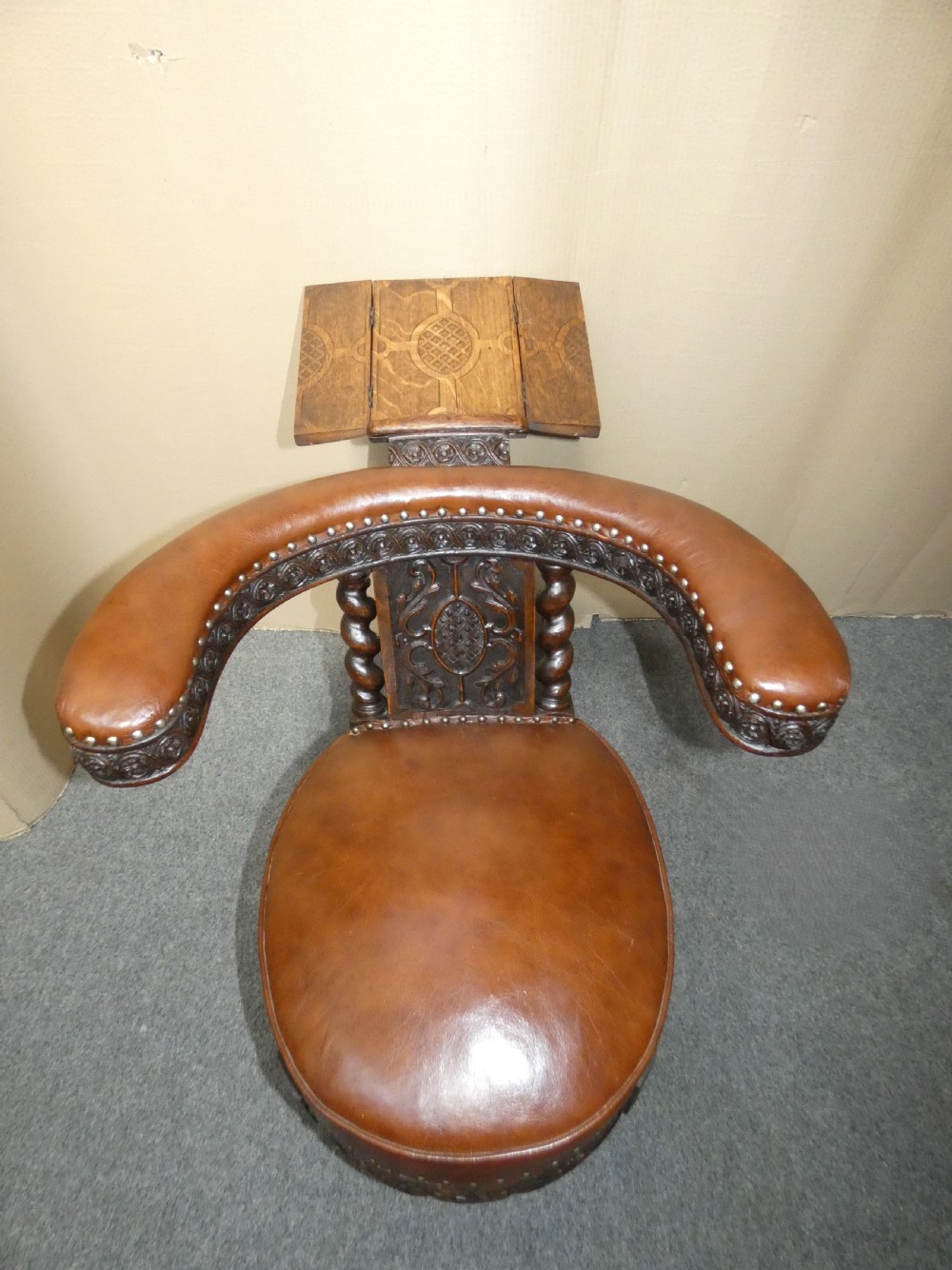 A carved dark oak cockfighting chair featuring tan leather upholstery with brass stud trimmings, a wide padded armrest, and barley twist support posts. View X
