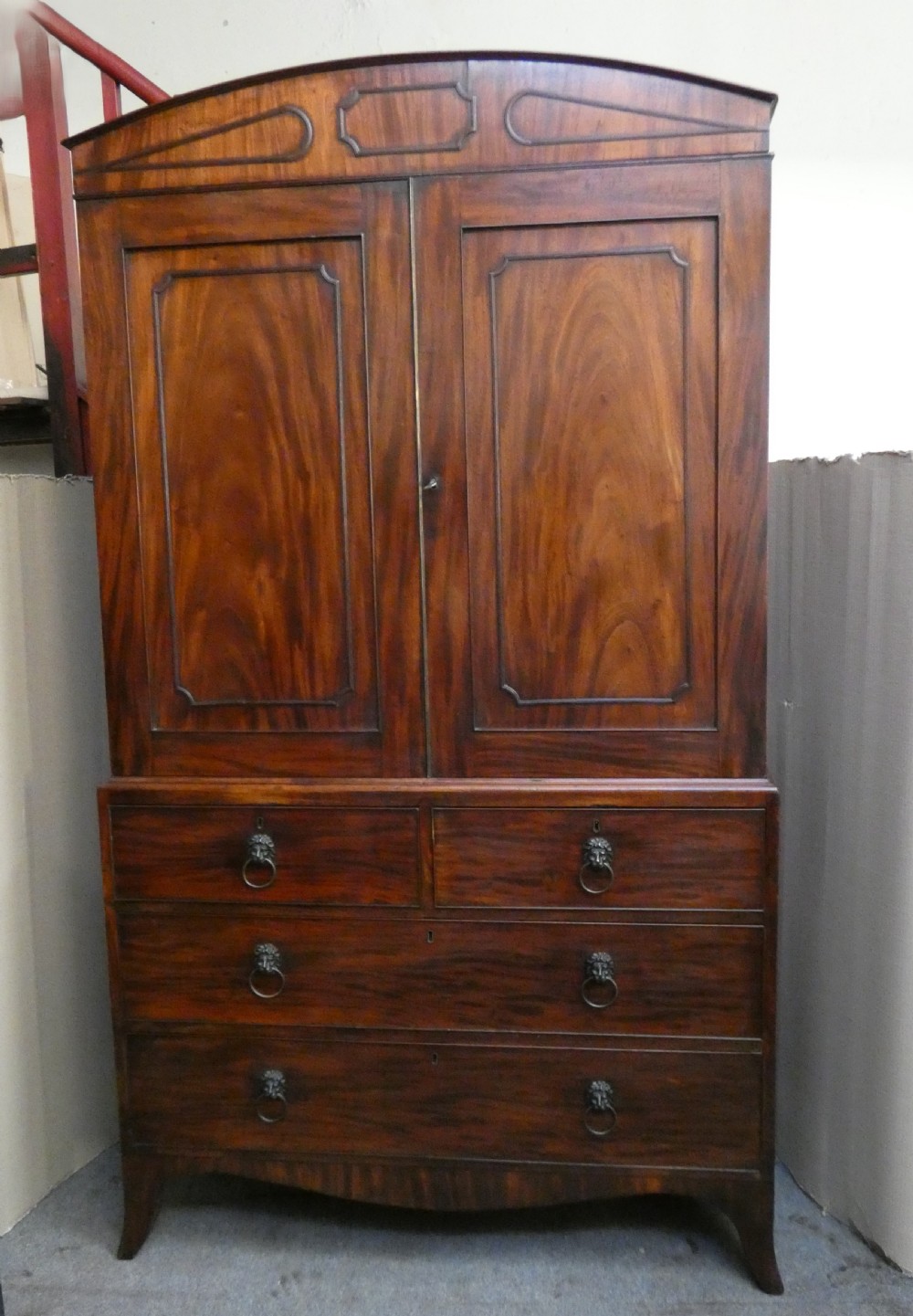 A Regency mahogany linen press featuring an arched pediment, twin panelled doors with geometric stringing, and a lower section with four drawers fitted with brass lion mask ring handles.