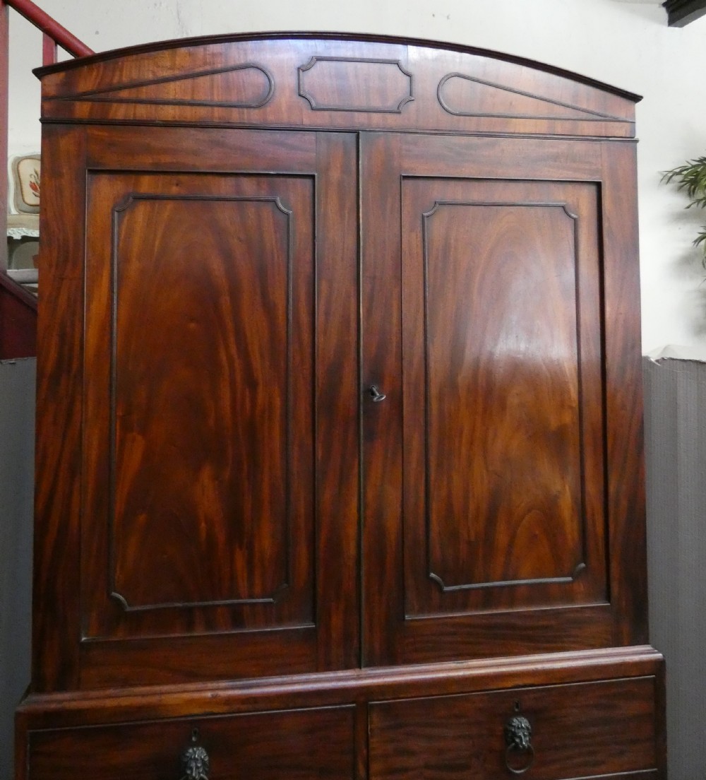 A Regency mahogany linen press featuring an arched pediment, twin panelled doors with geometric stringing, and a lower section with four drawers fitted with brass lion mask ring handles. View II