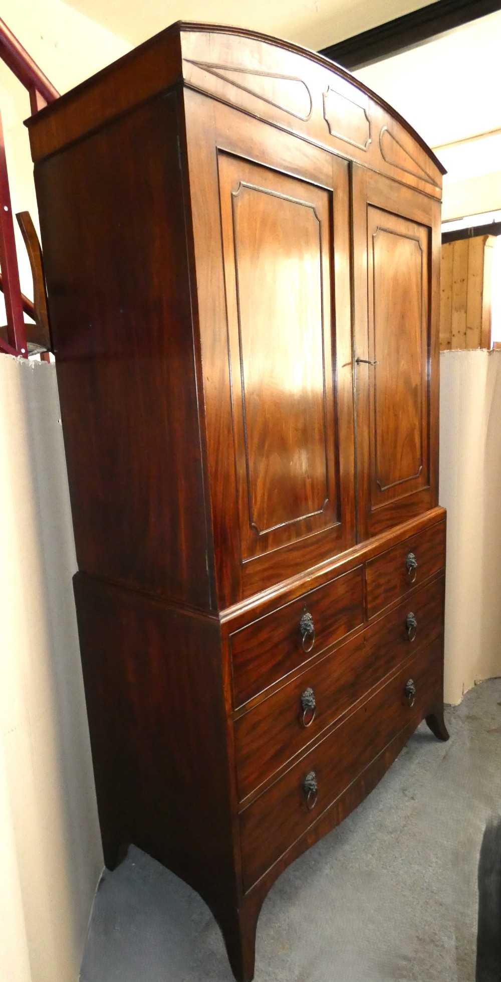 A Regency mahogany linen press featuring an arched pediment, twin panelled doors with geometric stringing, and a lower section with four drawers fitted with brass lion mask ring handles. View III
