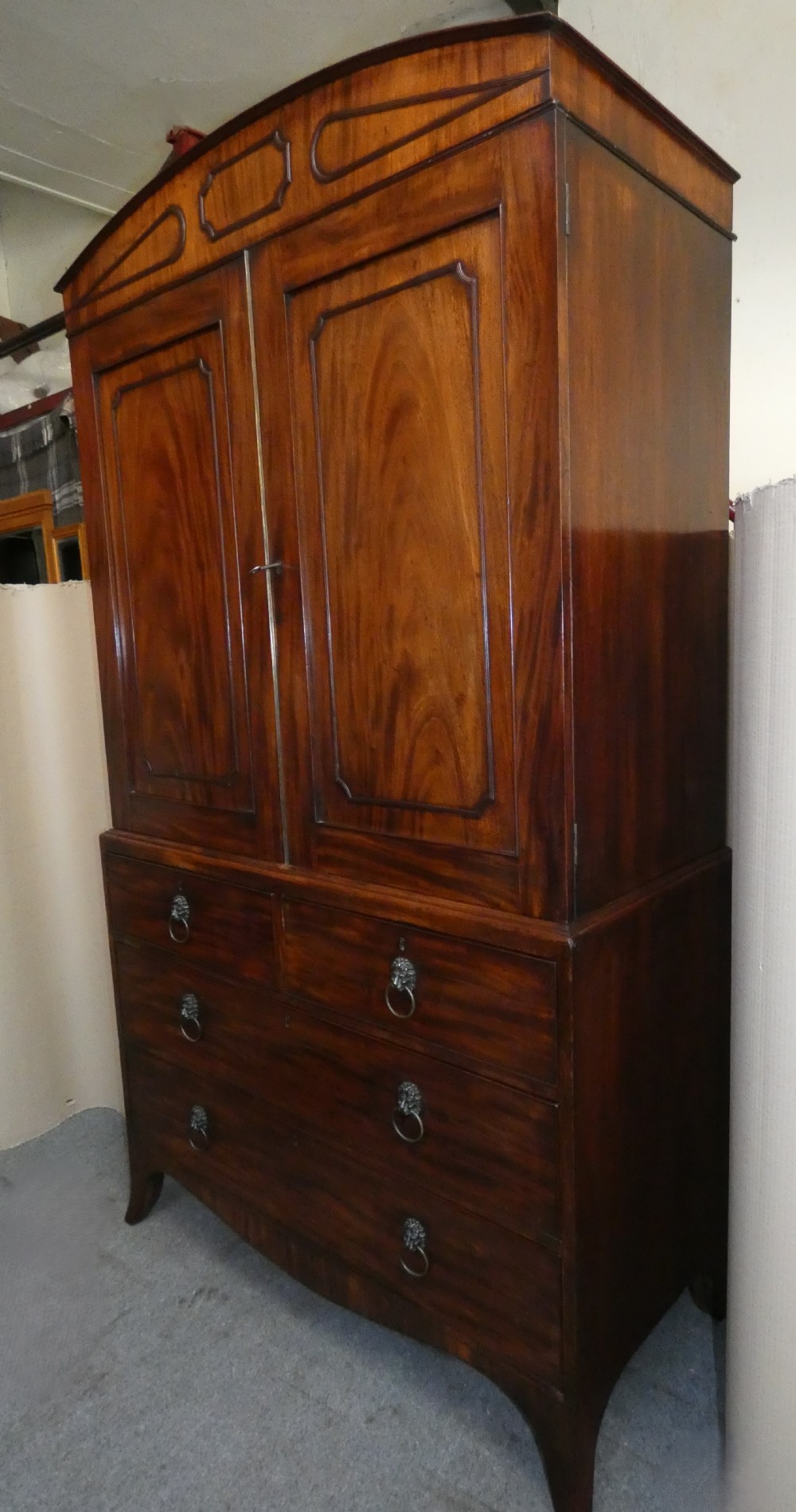 A Regency mahogany linen press featuring an arched pediment, twin panelled doors with geometric stringing, and a lower section with four drawers fitted with brass lion mask ring handles. View V