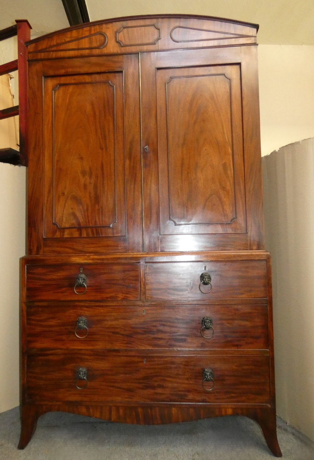 A Regency mahogany linen press featuring an arched pediment, twin panelled doors with geometric stringing, and a lower section with four drawers fitted with brass lion mask ring handles. View VII