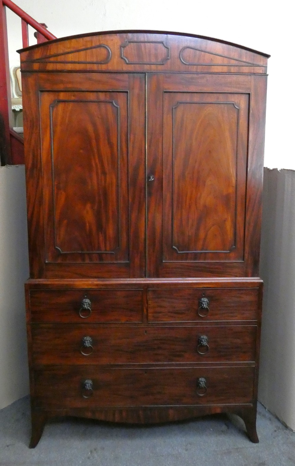 A Regency mahogany linen press featuring an arched pediment, twin panelled doors with geometric stringing, and a lower section with four drawers fitted with brass lion mask ring handles. View VIII