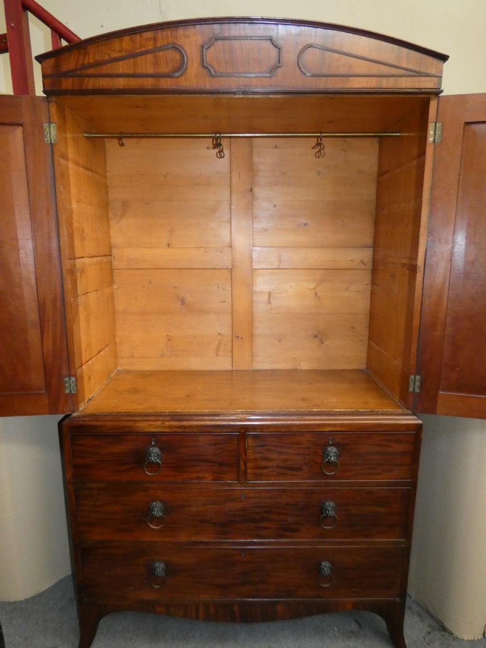 A Regency mahogany linen press featuring an arched pediment, twin panelled doors with geometric stringing, and a lower section with four drawers fitted with brass lion mask ring handles. View IX