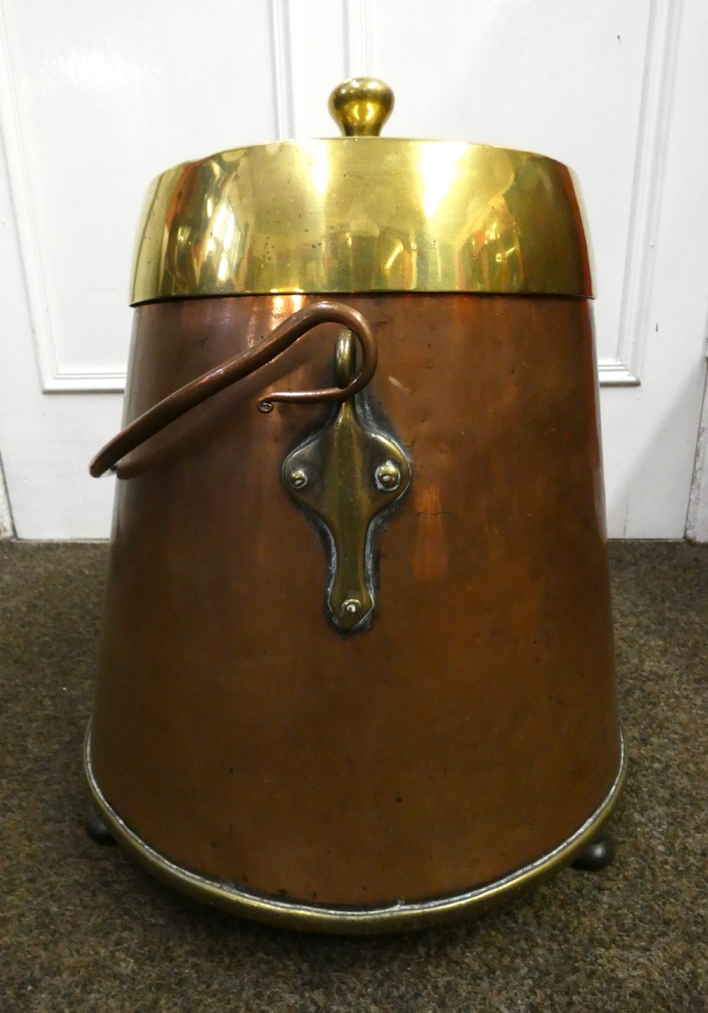 A tapered cylindrical copper coal bin featuring a polished brass collar and top finial, a copper swing handle, and decorative brass mounts.