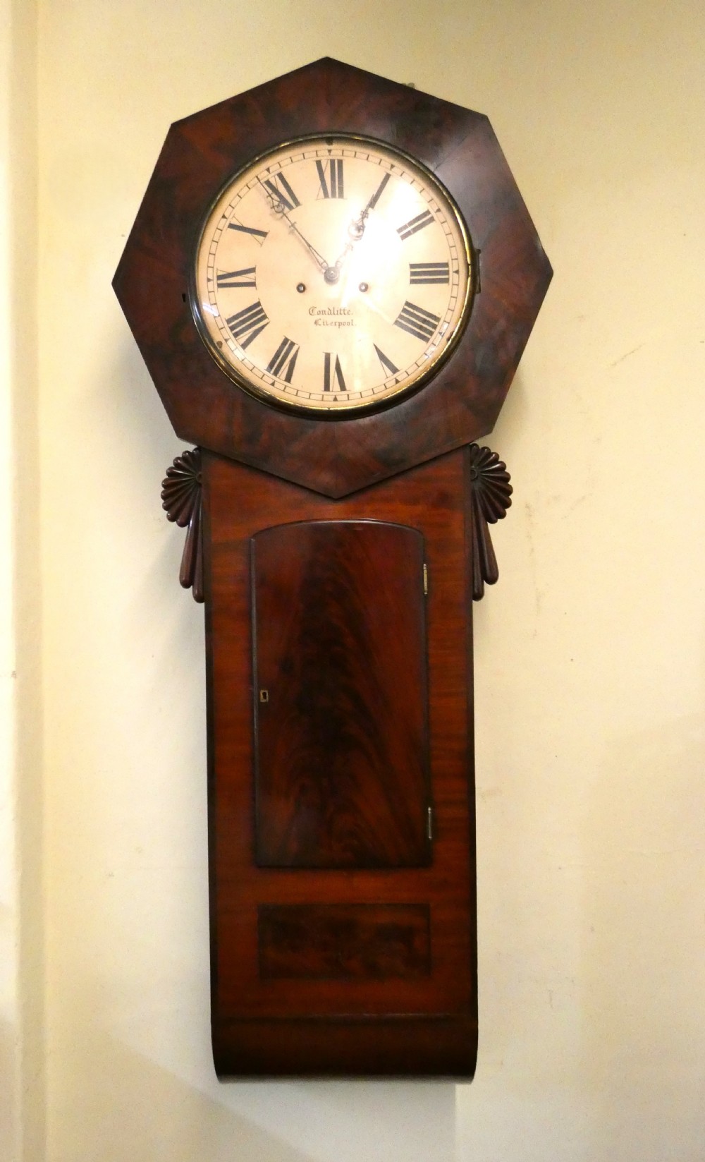 An antique mahogany tavern wall clock featuring an octagonal dial surround, carved side ears, and a long rectangular trunk with a flame mahogany door.